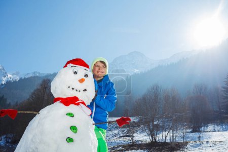 Téléchargez les photos : Beau jeune garçon en tenue de sport d'hiver stand avec bonhomme de neige fraîchement construit dans le chapeau de Père Noël et écharpe rouge à l'extérieur - en image libre de droit