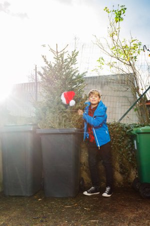 Foto de Niño joven tirar árbol de Navidad en el cubo de basura en el estacionamiento con sombrero de Santa, imagen conceptual que representa el final de las vacaciones - Imagen libre de derechos