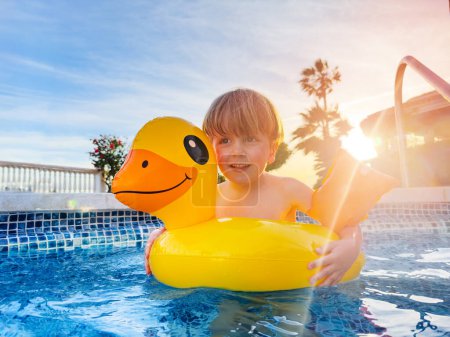 Foto de Niño en boya de pato y flotadores amarillos disfrutando de la piscina riendo con el atardecer soleado fondo iluminado - Imagen libre de derechos