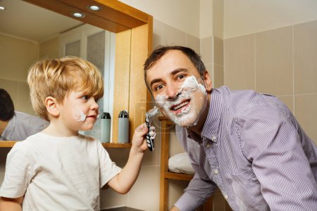 Téléchargez les photos : Petit gamin blond aidant son père souriant avec un visage mousseux pour se raser à l'aide d'un rasoir dans la salle de bain du matin - en image libre de droit
