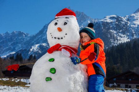 Photo for Handsome little boy in winter sport outfit play with snowman in Santa hat outside - Royalty Free Image