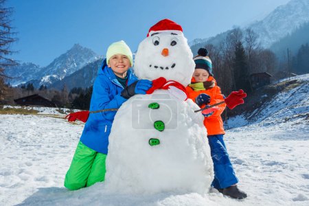 Téléchargez les photos : Portrait de deux garçons heureux frères en tenue de sport d'hiver jouer avec bonhomme de neige dans le chapeau du Père Noël et écharpe à l'extérieur - en image libre de droit