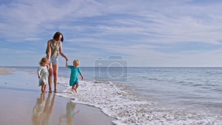Téléchargez les photos : Maman et deux petits enfants garçon, fille marcher sur la mer plage de sable jouer avec les vagues pendant les vacances d'été - en image libre de droit