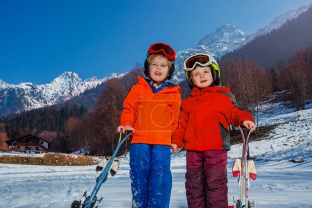 Foto de Primer plano de dos lindos niños pequeños en su primer puesto de vacaciones alpino con cielos usan cascos y máscara de esquí en la nieve sobre la montaña - Imagen libre de derechos