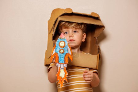 Téléchargez les photos : Serieux beau garçon jouer avec fusée en papier portant un casque d'astronaute en carton rêvant pour l'exploration spatiale - en image libre de droit