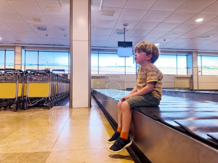 Photo for Handsome small boy sit and wait on the baggage conveyor belt waiting for checked in suitcase - Royalty Free Image