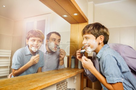 Téléchargez les photos : Papa et le fils s'amusent dans la salle de bain à se raser ensemble en regardant dans le miroir souriant portant chemise - en image libre de droit