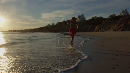 Foto de Niño en pantalones cortos de natación corren en la playa del océano salpicando en olas de agua bajo el sol naranja durante el atardecer en Francia, Bretagne - Imagen libre de derechos