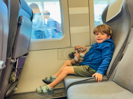Foto de Pequeño niño sentarse cerca de la ventana en el tren de alta velocidad abrazando peluche juguete con la cara feliz sonriente - Imagen libre de derechos
