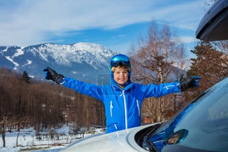 Téléchargez les photos : Heureux jeune garçon debout levant les mains souriant et porter un masque alpin avec casque par la voiture est arrivé pour les vacances de ski d'hiver - en image libre de droit