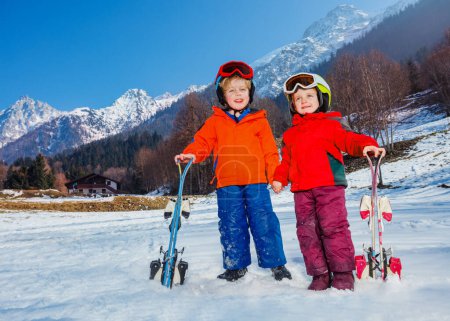 Foto de Dos niños felices en su primer puesto de vacaciones alpino con cielos usan cascos y máscara de esquí en la nieve sobre la montaña - Imagen libre de derechos