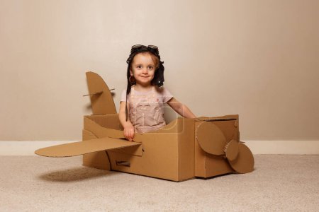 Foto de Hermosa chica sentarse en avión de cartón hecho a sí mismo con alas y hélice con sombrero piloto, gafas pretenden volar - Imagen libre de derechos