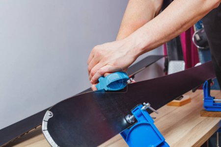 Téléchargez les photos : Homme dans un atelier tenir des skis sur des vises, utiliser un outil d'affûtage pour affûter les bords de ski de la rouille - en image libre de droit