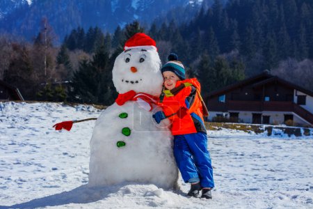 Photo for Portrait of a happy little boy in winter sport outfit play with snowman in Santa hat outside - Royalty Free Image