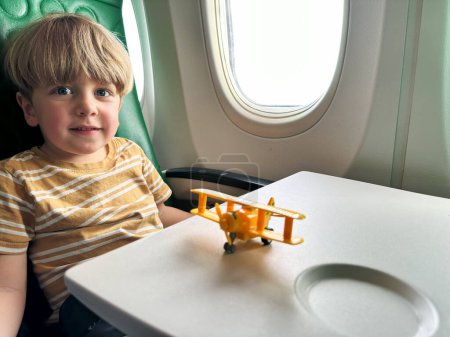 Foto de Guapo niño se sienta cerca de la ventana porthole en el avión jugando con juguete avión y sonrisa en la mesa - Imagen libre de derechos