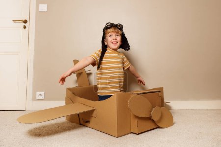 Photo for Happy handsome boy sit in self made cardboard plane with wings and propeller wearing pilot hat, glasses pretend to fly - Royalty Free Image