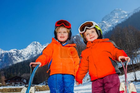 Photo for Close-up of two happy little kids on their first alpine vacation stand with skies wear helmets and skiing mask in snow over mountain - Royalty Free Image