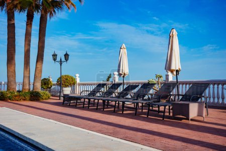 Téléchargez les photos : Chaise longue et parasols près de la piscine avec de grands palmiers sur le ciel bleu - en image libre de droit