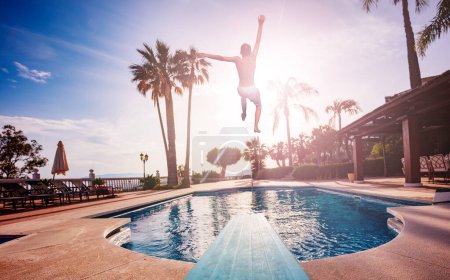 Téléchargez les photos : Garçon sauter de planche de plongée à la piscine, chaises longues et parasols éclairés avec la lumière du coucher de soleil chaud passant par les feuilles de palmier - en image libre de droit