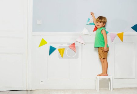 Photo for Little blond boy look at camera and put colorful party pennants chain, garland with flags on a wall in living room - Royalty Free Image