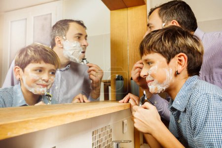Téléchargez les photos : Gros plan d'un père et le fils s'amusent dans la salle de bain se rasant ensemble en regardant dans le miroir souriant portant chemise - en image libre de droit
