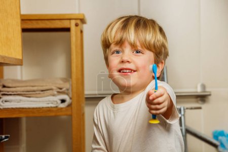 Foto de Retrato de un niño rubio cepillándose los dientes sosteniendo el cepillo de dientes en la mano y sonriendo a la cámara - Imagen libre de derechos