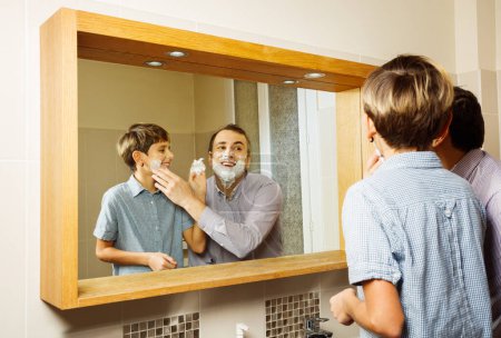 Téléchargez les photos : Père avec enfant pendant le rasage de liaison drôle et moussant, l'un l'autre dans la salle de bain, souriant, vue à travers le miroir - en image libre de droit