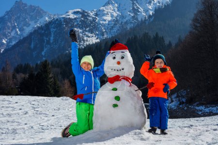 Photo for Two happy kids boys brothers in winter sport outfit play with snowman in Santa hat outside over mountains on background - Royalty Free Image
