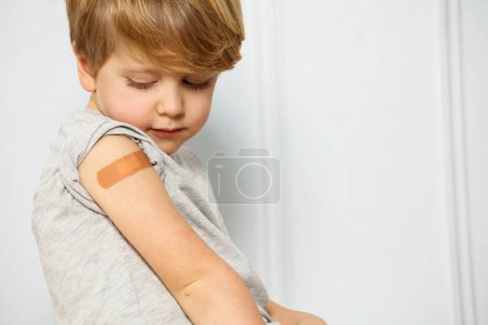 Photo for Handsome young boy with blond hair, who has a band-aid on shoulder and is looking at his hand scratch - Royalty Free Image