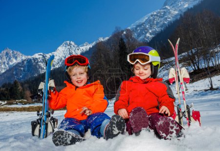 Photo for Two happy little kids on their first alpine vacation sit with skies wear helmets and skiing mask in snow over mountain - Royalty Free Image