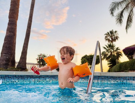 Photo for Joyful boy in arm bands enjoying sunset swimming pool having fun laughing and splashing - Royalty Free Image