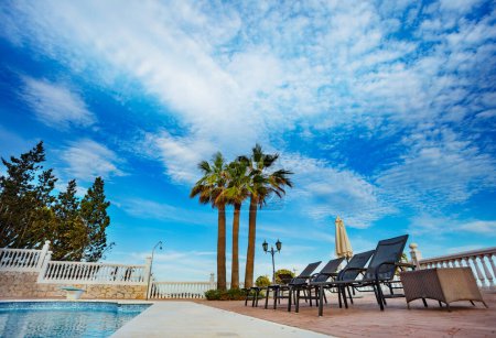 Photo for Typical vacation setup chaise longs near the pool and three big south palms over clear blue sky - Royalty Free Image