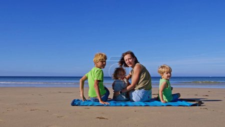 Foto de Madre con tres niños, niños y niñas se sientan en la alfombra de la playa en el océano volviendo sonriente - Imagen libre de derechos