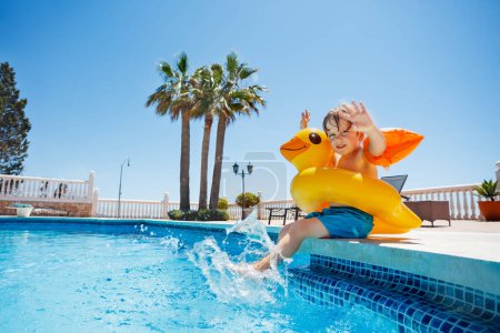 Boy at resort pool enjoys summer, splashing in buoy and floaties, lift hands over palm trees, blue sky
