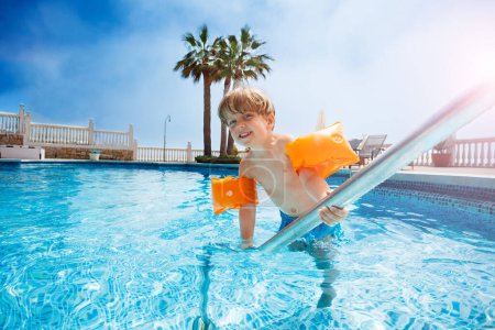 Photo for Happy boy in arm bands entering clear water of the pool smiling looking in camera - Royalty Free Image