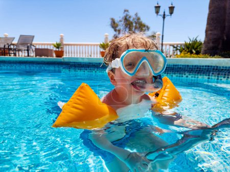 Photo for Happy blond boy in the swimming mask and arm bands floating in pool on a sunny weather - Royalty Free Image