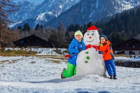 Téléchargez les photos : Deux enfants heureux garçons frères en tenue de sport d'hiver jouer avec bonhomme de neige dans le chapeau de Père Noël à l'extérieur sur les montagnes sur fond - en image libre de droit