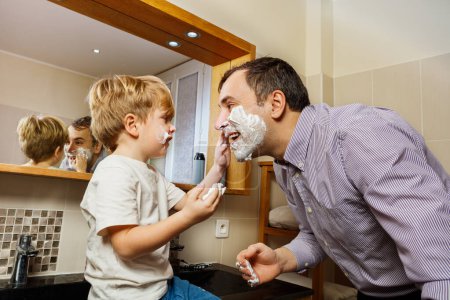 Téléchargez les photos : Drôle de famille moments petit enfant moussant papa visage souriant dans la salle de bain du matin - en image libre de droit