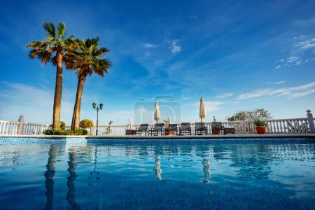 Téléchargez les photos : Scène de vacances relaxante sous un ciel sans nuages avec chaises longues autour d'une piscine calme, et trois palmiers - en image libre de droit
