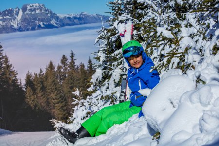 Foto de Niño feliz en casco de esquí con googles y cielos alpinos sonrisa sentado en las montañas de nieve sobre abetos nevados - Imagen libre de derechos