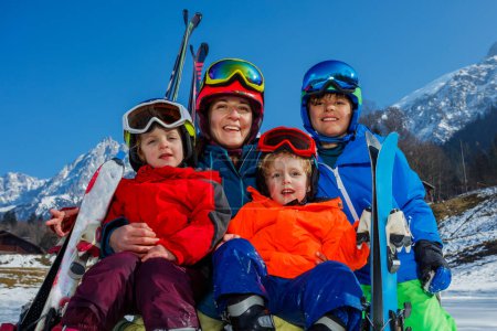 Téléchargez les photos : Gros plan portrait de famille de quatre personnes assises dans la neige, mère, garçon et deux petits enfants vêtus de casques de tenue de sport, riant en souriant en profitant de la belle vue sur la montagne - en image libre de droit