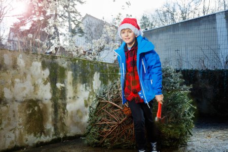 Téléchargez les photos : Image en gros plan d'un jeune garçon heureux avec une hache pour couper les arbres à la main dans le chapeau de Père Noël ramener à la maison arbre de Noël pour la célébration du Nouvel An tenant tronc et souriant - en image libre de droit