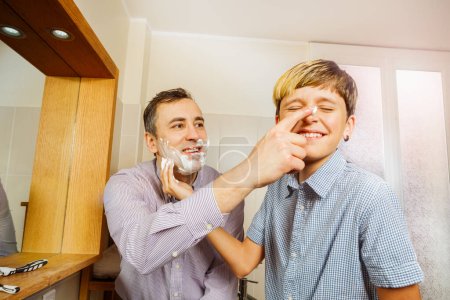 Téléchargez les photos : Souriant papa a mis de la mousse à raser sur son nez d'enfant en s'amusant à se préparer à se raser dans la salle de bain ensoleillée - en image libre de droit