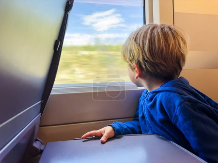 Téléchargez les photos : Petit garçon assis et regardant la fenêtre à l'intérieur du train à grande vitesse regardant le paysage rural - en image libre de droit
