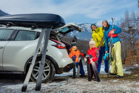 Photo for Family with father mother and two adorable little kids standing joking holding ski by the open car trunk - Royalty Free Image
