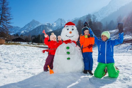 Téléchargez les photos : Enfants heureux, les garçons et une fille construire bonhomme de neige jouer à l'extérieur dans la région montagneuse posant les mains levantes - en image libre de droit