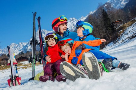 Fun on first ski alpine vacation family of mom and three kids boys with girls sit hugging laughing in snow wear helmets sport outfit masks