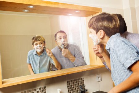 Téléchargez les photos : Papa et adolescent gosse rasage ensemble regarder miroir, souriant dans salle de bain ensoleillée - en image libre de droit