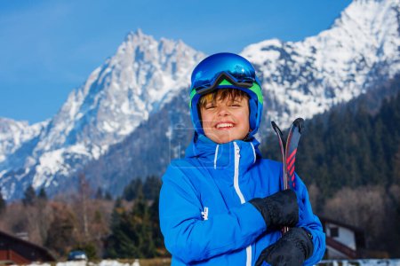 Photo for Handsome boy, dressed in an alpine ski outfit, a mask and helmet, stands over snow covered mountains grinning with wide smile - Royalty Free Image