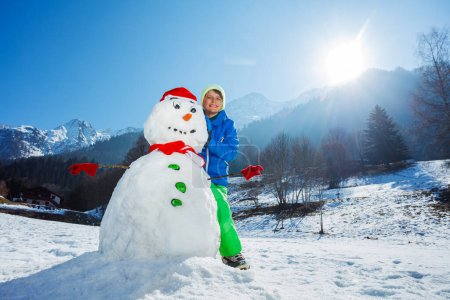 Téléchargez les photos : Portrait d'un jeune garçon heureux en tenue de sport d'hiver stand avec bonhomme de neige fraîchement construit dans le chapeau du Père Noël et écharpe rouge à l'extérieur - en image libre de droit